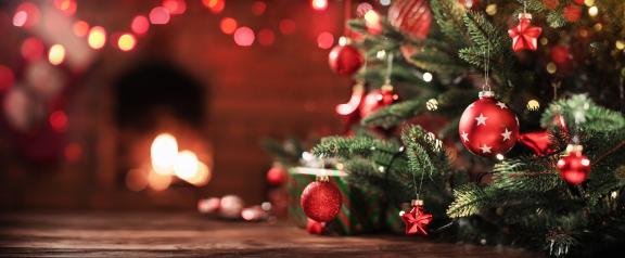 Christmas tree with red ornaments in front of a glowing fireplace and blurred festive lights