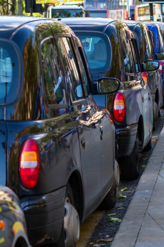 A row of black London-style taxis parked closely along a curb on a city street. The vehicles are aligned in a neat line, with their rear lights and side mirrors visible.