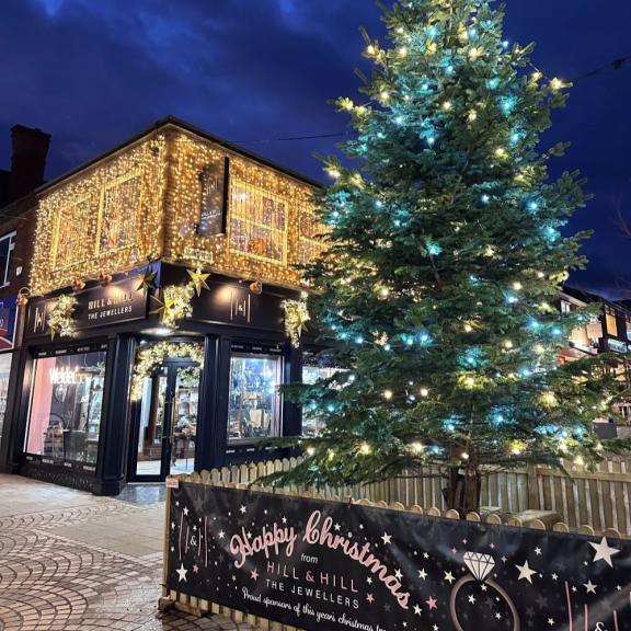 A large outdoor Christmas tree decorated with white and blue lights stands next to Hill & Hill The Jewellers, a shop with its exterior covered in golden fairy lights