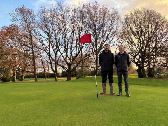 Two people standing on a golf green beside a red flag, with bare trees and a sunset sky in the background.