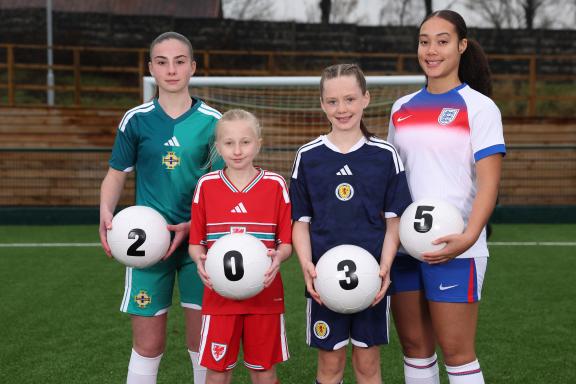 Four football players standing on an artificial turf pitch, each wearing a different national team kit: green for Northern Ireland, red for Wales, navy for Scotland, and white for England. Each player is holding a white football with a black number, collectively forming “2 0 3 5.” A goalpost and wooden fencing are visible in the background.