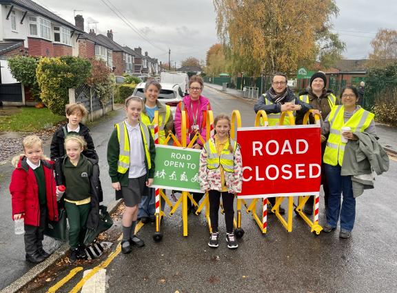 Group of people wearing high-visibility jackets standing on a residential street behind yellow barriers with two signs: one green sign reading “ROAD OPEN TO” with symbols for walking and cycling, and one red sign reading “ROAD CLOSED.” Houses line the street, and autumn trees are visible in the background under a cloudy sky.