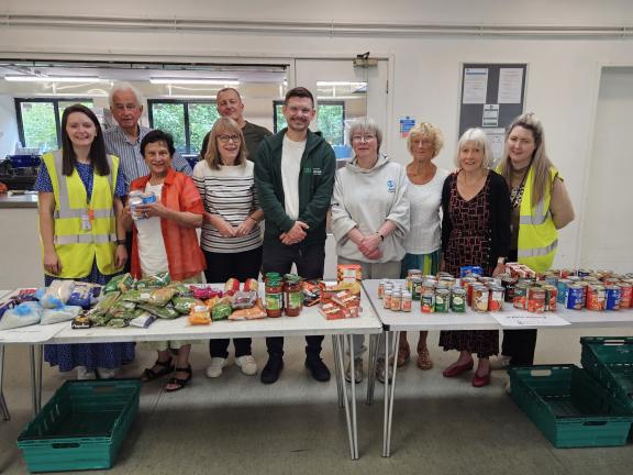 A group of volunteers standing behind two tables filled with assorted food items, including fresh vegetables, jars, and canned goods, in a community hall setting.