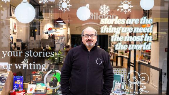 Photo shows the storefront of “Share Your Story” in Altrincham, featuring staff member Simon standing in front of the glass window.