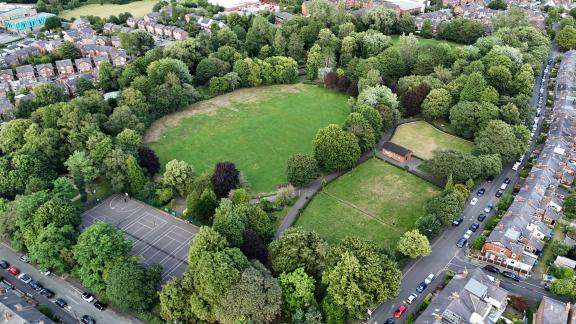 Aerial view of Stamford Park showing a large grassy field surrounded by dense trees, with a sports court, smaller lawn areas, and nearby residential streets lined with houses and parked cars.