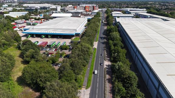Aerial view of Trafford Park with large warehouses, colorful trucks, and a tree-lined road.