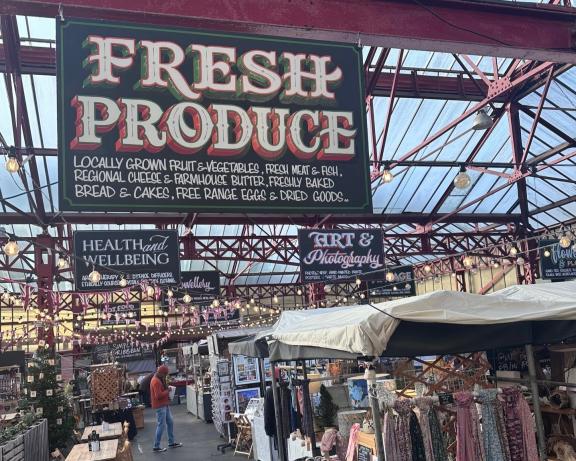 Indoor market with red metal beams and a glass roof, featuring a large “Fresh Produce” sign advertising locally grown goods