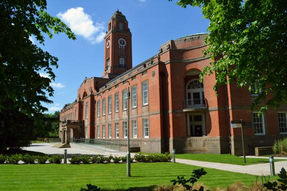 Trafford Town Hall, a historic red-brick building with a central clock tower, tall arched windows, and ornate stone detailing, surrounded by green lawns, trees and a paved walkway under a clear blue sky.
