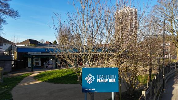 Exterior of the Trafford Family Hub in Stretford on a sunny day, with trees and a blue sign at the entrance.