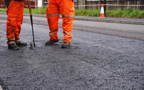 Two road workers in bright orange safety gear resurfacing a street with fresh tarmac.