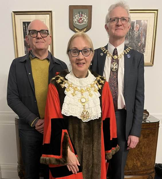 Three people stand indoors, with one in front wearing ceremonial mayoral robes and chains, and two others behind dressed in smart clothing, posing for a formal photograph in an office setting.