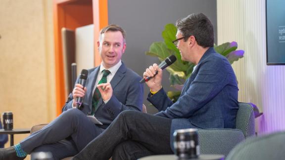 Two people sit on stage-facing chairs, each holding a microphone while engaged in conversation. 