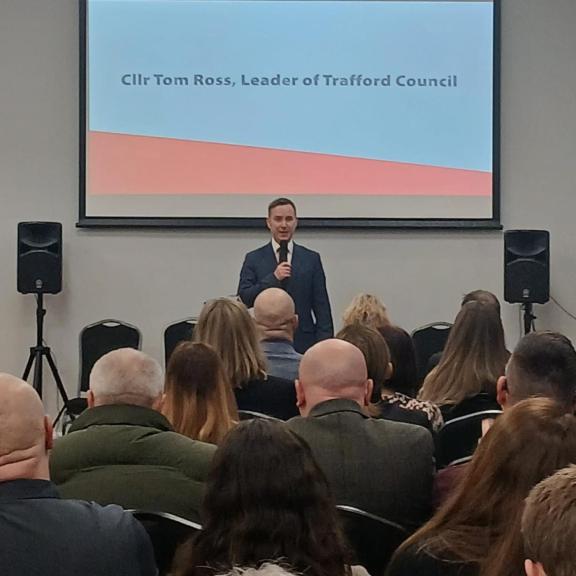 Cllr Tom Ross stands at the front of a conference room holding a microphone, addressing an audience seated in rows