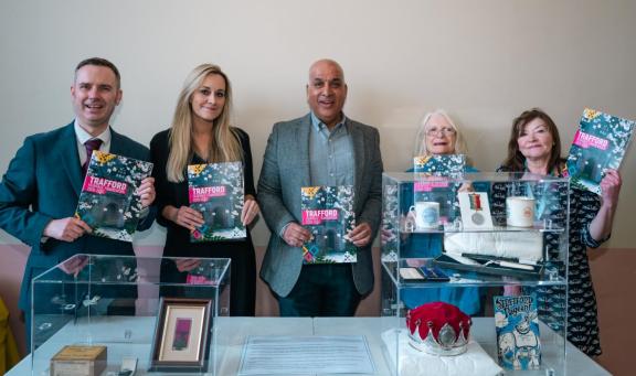 A group of councillors stand behind a display table holding copies of a publication titled “Trafford Heritage Framework”. The table in front of them contains several glass display cases with various heritage items, including framed documents, small objects, textiles, medals, and a red ceremonial-style item. 