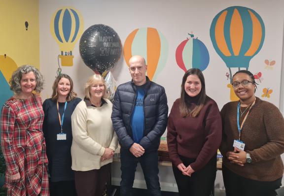 A group of six adults stand together in a brightly decorated room with colourful hot‑air balloon wall art and birthday balloons behind them.