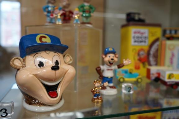 Close-up of vintage Kellogg’s memorabilia displayed in a glass cabinet, including a smiling cartoon character head jar wearing a blue cap with a “C,” small figurines, and retro cereal packaging in the background.