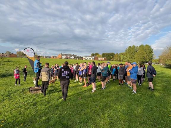 A large group of people gathered on a grassy field for a community running event, listening to a speaker standing on a small platform beside a flag banner. 