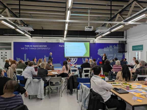 People seated at round tables in a large indoor event space watch a presentation projected on a screen, while two presenters stand at the front