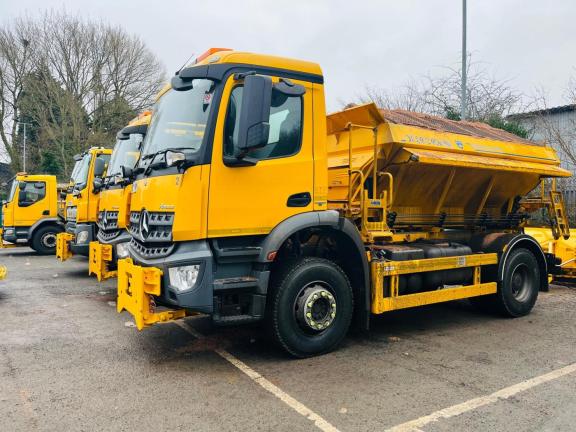 Row of bright yellow grit-spreading lorries parked in a lot on an overcast day