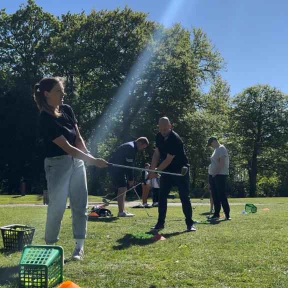 People practicing golf swings on a grassy driving range under bright sunlight, with several baskets of golf balls on the ground and a line of trees in the background.