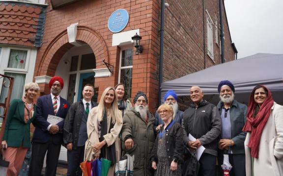 Group of people standing outside a red brick house beneath a blue commemorative plaque. The plaque, mounted above an arched doorway, honors Gyani Sundar Singh Sagar.