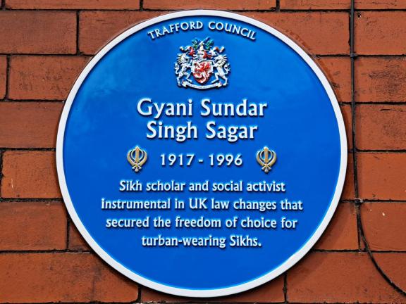 Blue circular commemorative plaque on a red brick wall reading “Trafford Council” with a crest above the name Gyani Sundar Singh Sagar (1917–1996). The plaque notes that he was a Sikh scholar and social activist instrumental in UK law changes securing freedom of choice for turban-wearing Sikhs.