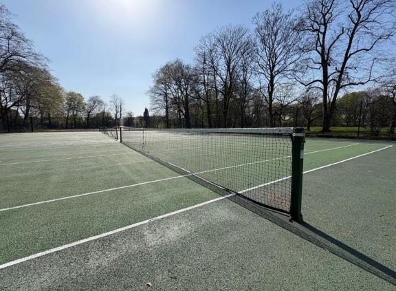 Empty outdoor tennis courts in Longford Park, viewed along the net stretching across the middle. The green hard surfaces are marked with white lines, and tall leafless trees line the background under a clear blue sky.
