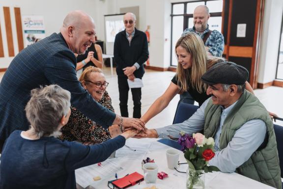 A group of adults gathered around a table indoors place their hands together in the center as part of a group activity. 