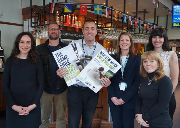 Group of six adults in a bar holding domestic abuse awareness posters, with international flags and glassware visible behind them. from left to right: Cllr Keleigh Glenton, Executive Member for Community Safety and Civil Contingencies; John Lloyd, GMP Licensing Officer for Trafford; Ben Plunkett, Manager of The Bishop Blaize Wetherspoons; Nicola Walton, Domestic Abuse Co-ordinator, Trafford Council; Cllr Jo Harding, Deputy Mayor of Trafford and Executive Lead for Violence Against Women and Girls; Samantha F
