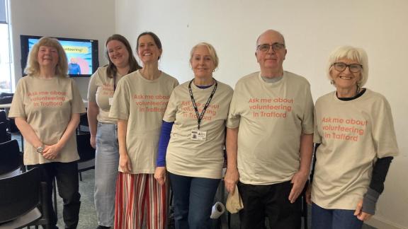 Six people stand side by side indoors wearing matching beige T‑shirts with the printed slogan “Ask me about volunteering in Trafford.” The group faces the camera in a room with chairs and a screen in the background displaying a volunteering-themed slide.