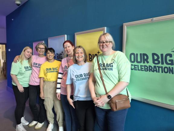 A group of seven people standing together indoors against a blue wall, wearing pastel T-shirts that read “Our Big Celebration,” with colorful framed panels behind them.