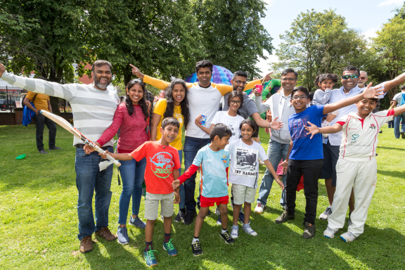 A group of adults and children celebrating at Trafford Live in 2023. Some ae dressed in cricket whites, while others are waving cricket bats around in celebration