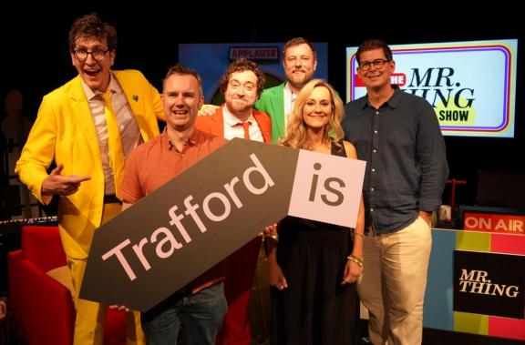 Six people, including the comedy group "Mr. Thing," pose together in a brightly lit theatre. They are dressed in vibrant colours. In the centre, they hold the same large "Trafford Is" arrow sign. A sign in the background reads "APPLAUSE".