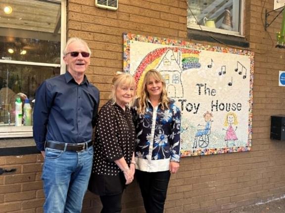Three adults stand outside a brick building beside a large decorative sign reading “The Toy House.” The sign features a rainbow, musical notes, a house illustration, and drawings of children. A window is visible on the left side of the building, and the group is posed in front of the sign.