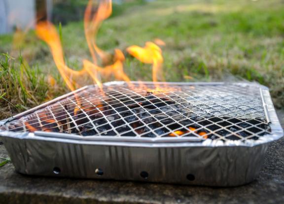 Disposable foil barbecue tray with a wire grill and glowing charcoal, producing visible flames outdoors on a stone surface beside a grassy lawn.