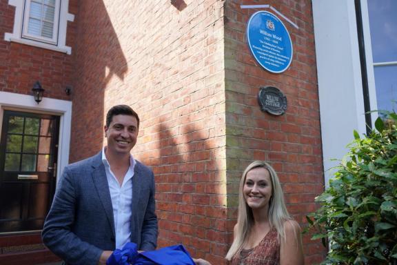 Two people stand outside a red-brick house beneath a blue Trafford Council plaque commemorating William Wood. The plaque is mounted on the corner wall above a sign reading “Willow Cottage,” with one person holding a blue unveiling cloth.