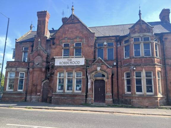 Front view of a large, disused red-brick Victorian building with Gothic architectural details, chimneys, bay windows, and a central entrance.