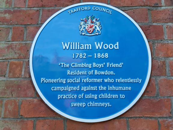 Blue commemorative plaque mounted on a red brick wall. The plaque, issued by Trafford Council, honours William Wood (1782-1868), known as “The Climbing Boys’ Friend,” a resident of Bowdon and pioneering social reformer who campaigned against the use of children as chimney sweeps. The Trafford Council crest appears at the top of the plaque.