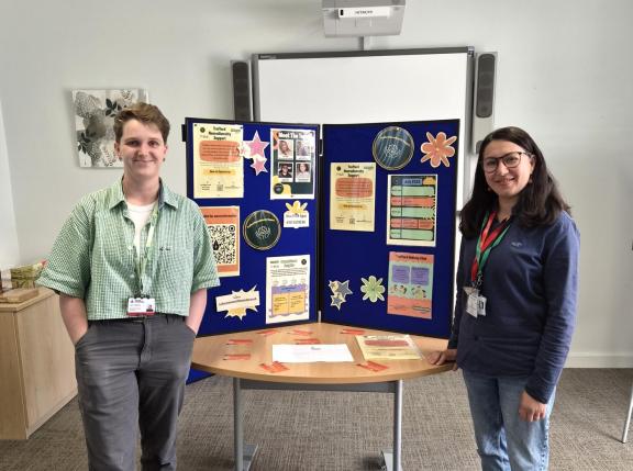 Two people wearing staff lanyards stand on either side of a table displaying information about Trafford Neurodiversity Support.