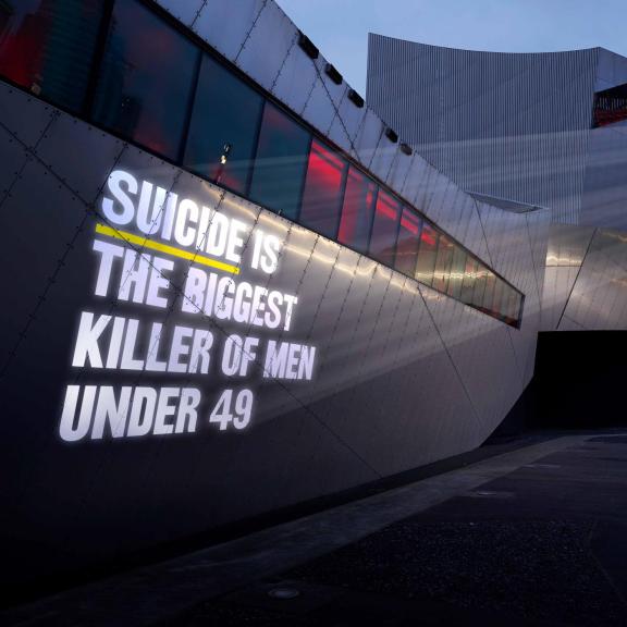 Large suicide awareness message projected onto the metallic exterior of the Imperial War Museum North at dusk. The illuminated text reads “Suicide is the biggest killer of men under 49”.