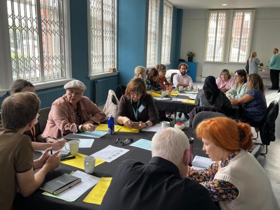 A community workshop taking place in a bright meeting room with large blue walls and tall windows fitted with security grilles. Around a dozen participants are seated at round tables covered with black tablecloths, discussing and completing worksheets.