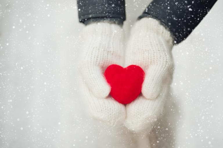 White knit gloves holding a small red heart in snowy weather