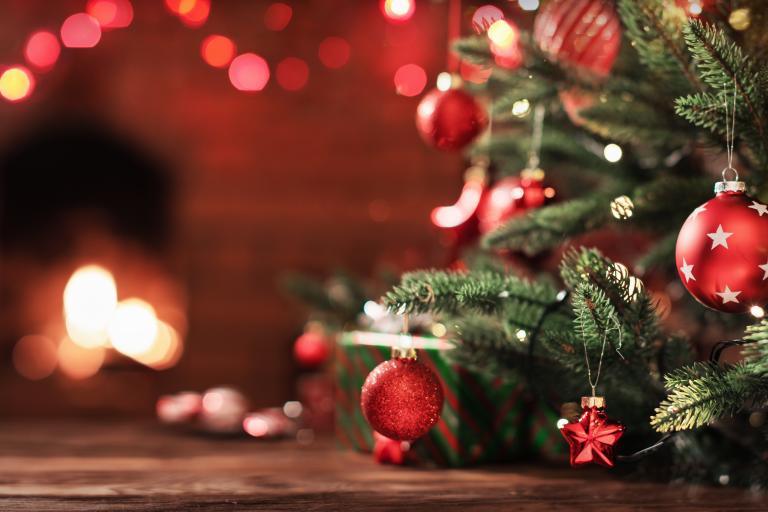 Christmas tree with red ornaments in front of a glowing fireplace and blurred festive lights