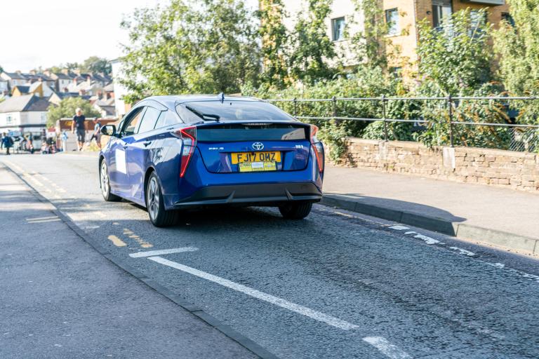 A blue Toyota Prius taxi driving on a suburban road with a double yellow line. The car has a yellow license plate and a taxi sign on the rear window. T