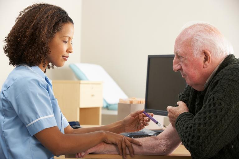 A healthcare professional in a blue uniform sits at a desk holding a syringe near an older person’s arm, preparing for an injection. A computer monitor and medical supplies are visible in the background.