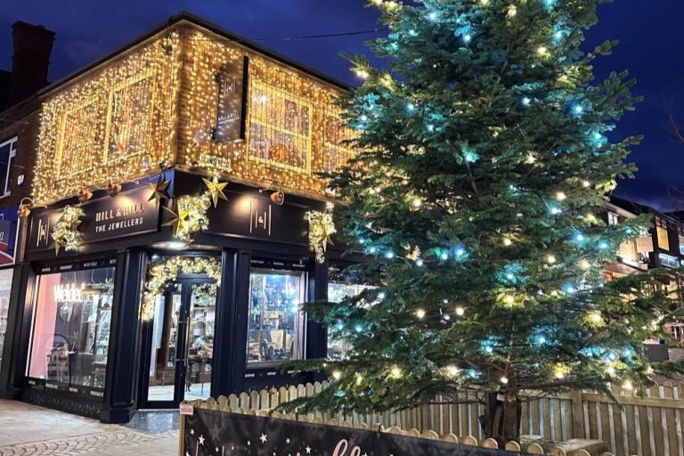 A large outdoor Christmas tree decorated with white and blue lights stands next to Hill & Hill The Jewellers, a shop with its exterior covered in golden fairy lights