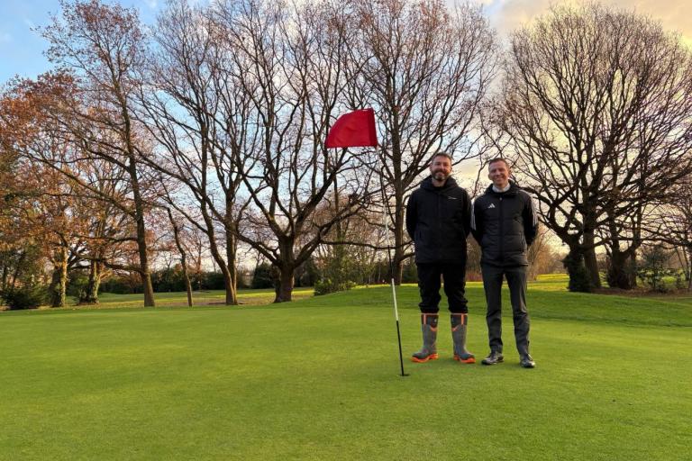 Two people standing on a golf green beside a red flag, with bare trees and a sunset sky in the background.