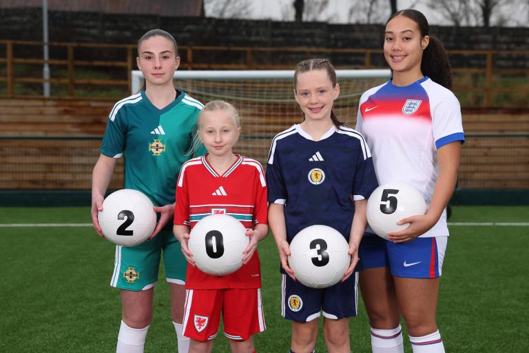 Four football players standing on an artificial turf pitch, each wearing a different national team kit: green for Northern Ireland, red for Wales, navy for Scotland, and white for England. Each player is holding a white football with a black number, collectively forming “2 0 3 5.” A goalpost and wooden fencing are visible in the background.