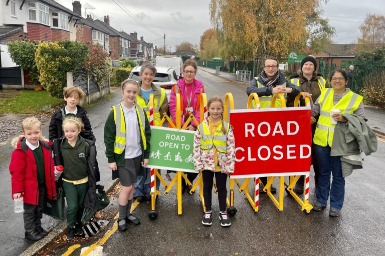 Group of people wearing high-visibility jackets standing on a residential street behind yellow barriers with two signs: one green sign reading “ROAD OPEN TO” with symbols for walking and cycling, and one red sign reading “ROAD CLOSED.” Houses line the street, and autumn trees are visible in the background under a cloudy sky.