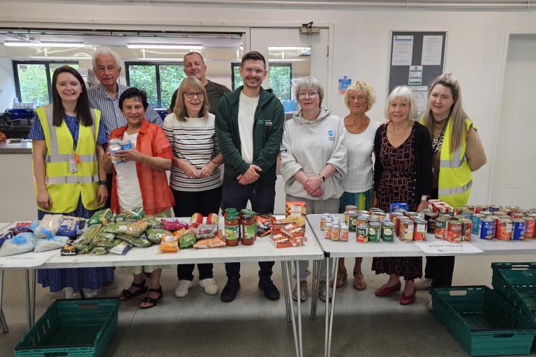 A group of volunteers standing behind two tables filled with assorted food items, including fresh vegetables, jars, and canned goods, in a community hall setting.